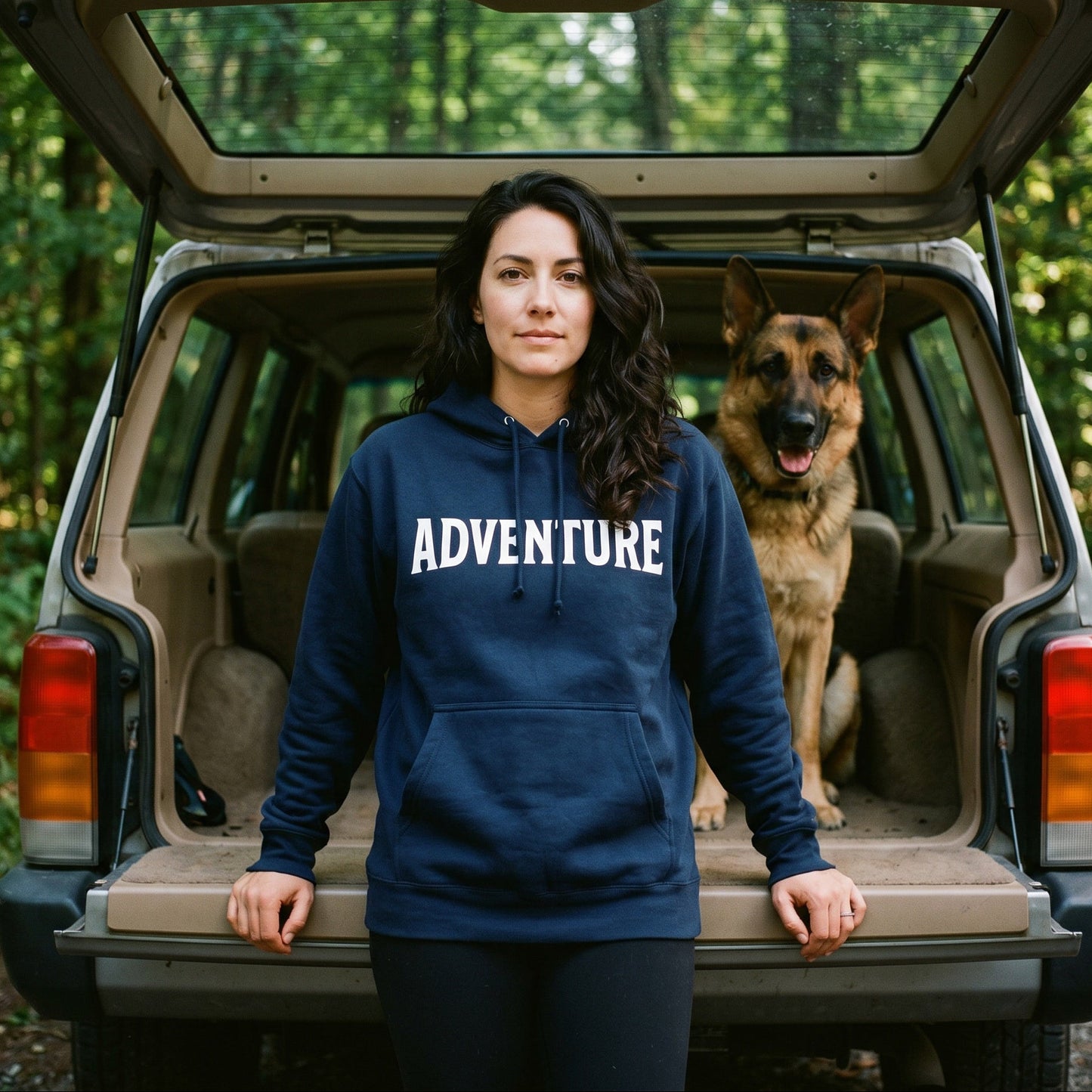 Woman wearing a hoodie with 'ADVENTURE' printed on it, standing next to a dog in an open car trunk surrounded by trees.